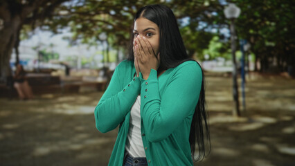 Naklejka premium Woman in green cardigan covers mouth with both hands in a sunlit outdoor park setting; astonishment.