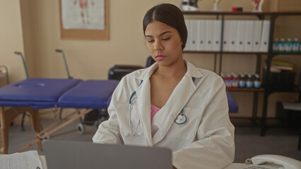 Latin woman doctor in white coat typing on laptop computer at clinic desk in building; concentration.