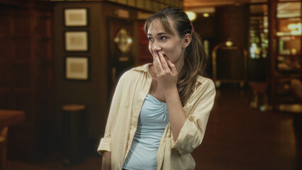 Young woman covering mouth with hand while smiling in a hotel lobby building with bell cart and framed art visible; surprise.
