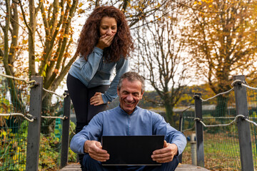 Happy couple laughing while watching a laptop video call on a bridge in an autumn park