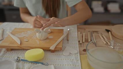 Girl sculpts wet clay on wooden table with hands in art studio, forming small pot beside pottery tools; creative passion.