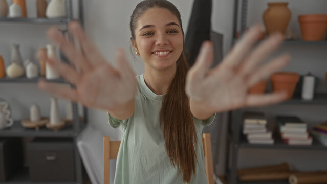 Teenage girl smiling extends wet clay smeared hands in sunlit pottery studio; creative expression and joy. - Powered by Adobe