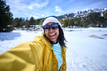 POV of African American young woman taking selfie with two hands with mobile in snowy mountains. Generation Z Latin beautiful female in yellow looking smiling at camera sunny day of winter holidays 