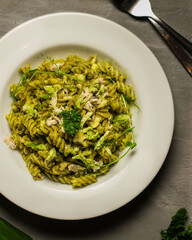 A close-up top view of creamy green pesto fusilli pasta served in a white plate. The dish is garnished with shredded chicken, fresh herbs, and greens, creating a vibrant and appetizing presentation. 
