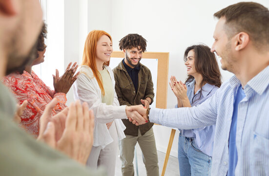 Smiling man and woman in casual wear shaking hands, reaching agreement, making deal, signing contract or greeting new employee with happy colleagues or groupmates congratulating and applauding around.
