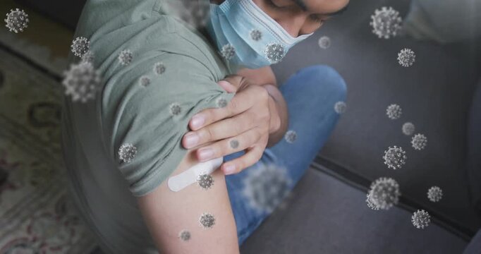 Man sitting on sofa lifting sleeve and checking bandage while virus graphics overlaying health