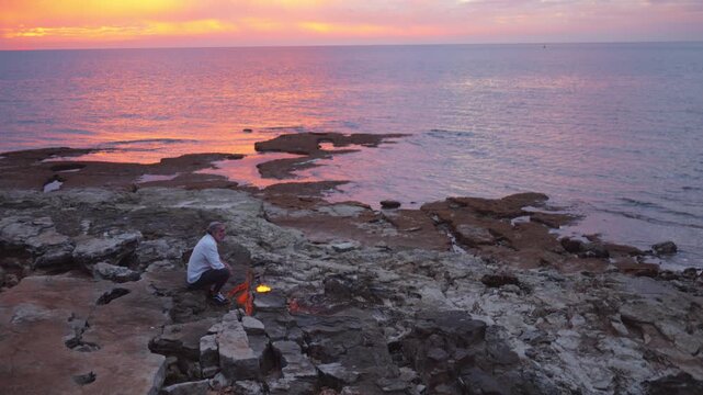A person sits on rocky terrain by the sea at sunset, gazing at a small fire. The serene ocean and vibrant sky evoke mindfulness and reflection, offering a tranquil escape from daily life.