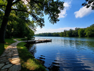 ** Scenic view of a calm lake with a wooden dock, framed by lush green trees and a blue sky dotted with fluffy clouds. A stone path winds along the shore. **