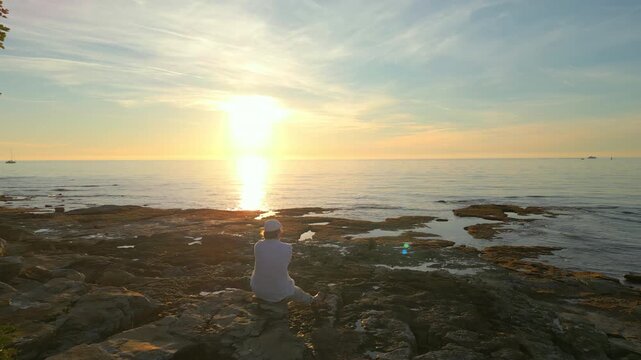 A solitary figure sits on rocky shores at sunset, gazing at the horizon. The serene seascape with gentle waves and distant sailboats evokes mindfulness and reflection.