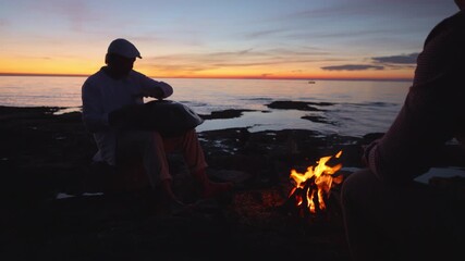 Two individuals sit by a campfire at sunset by the sea, reflecting quietly. The serene setting promotes mindfulness and escape, emphasizing mental wellbeing and nature connection.