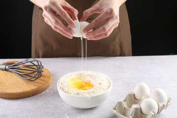 Woman breaking egg into bowl with flour at grey textured table against black background, closeup