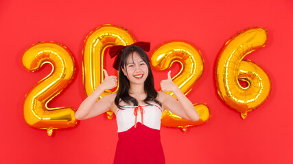 Asian young cheerful beautiful woman wearing red dress posing with golden balloon shape number figure 2026 on red background smiling giving thumbs up Happy New Year celebration festive holiday concept