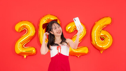 Asian young cheerful woman smiling wearing red dress video call greeting mobile phone posing with golden balloon shape number figure 2026 on red background, Happy New Year celebration festive holiday.
