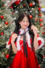 young asian woman in a red santa dress posing among decorated christmas trees with warm festive lights and red ornaments, captured with soft bokeh for holiday-themed