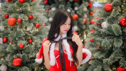 young asian woman in a red santa dress posing among decorated christmas trees with warm festive lights and red ornaments, captured with soft bokeh for holiday-themed