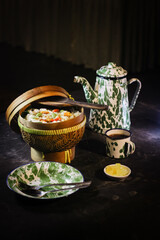 A rustic meal setup with a woven basket of seasoned rice, a vintage enamel plate with cutlery, and a cup of black coffee on a dark table. Warm lighting creates a homely, traditional Indonesian dining.