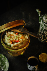 A rustic meal setup with a woven basket of seasoned rice, a vintage enamel plate with cutlery, and a cup of black coffee on a dark table. Warm lighting creates a homely, traditional Indonesian dining.