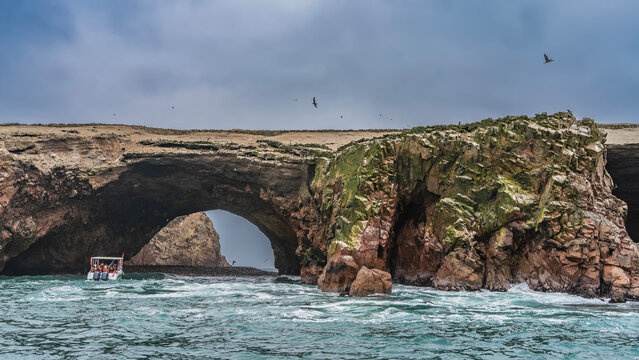 Picturesque cliffs in the ocean. A tourist ship sails to a natural stone arch. The waves of the turquoise sea beat against the rocks. Birds are flying in the sky. Peru. Paracas. Ballestas Islands 