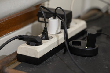 Close-up of a power strip with various chargers. A camera battery charger shows a green light, indicating a full charge.