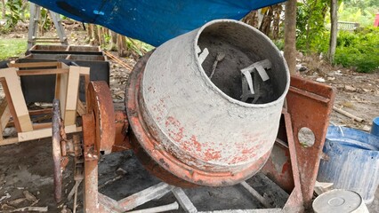 A worn concrete mixer machine in an outdoor construction area, surrounded by tools and materials under a blue tarp. This photo is perfect for content related to construction work, industry, building e