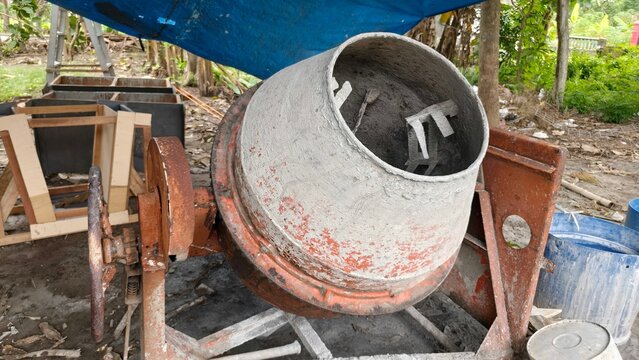 A worn concrete mixer machine in an outdoor construction area, surrounded by tools and materials under a blue tarp. This photo is perfect for content related to construction work, industry, building e