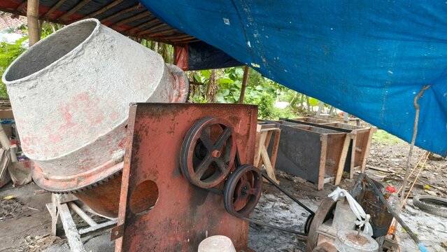 A worn concrete mixer machine in an outdoor construction area, surrounded by tools and materials under a blue tarp. This photo is perfect for content related to construction work, industry, building e