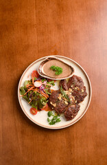 ​A gourmet dish of grilled steak slices topped with sauce, served alongside a side salad on a white plate, set on a warm wooden dining table.