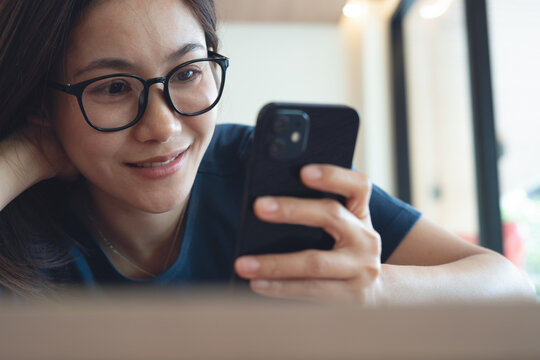 Close up, smiling young adult asian woman sitting at table at home using smartphone and watching at the screen, domestic life. Happy female hipster watching video viral on mobile phone