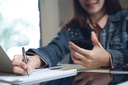 Young asian business woman using smartphone and laptop computer, working at office. Businesswoman doing online research and taking note on notepad, business planning. Student online studying, close up
