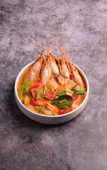 A vibrant and spicy bowl of Thai Tom Yum Goong soup, overflowing with large, fresh river prawns and colorful vegetables, set against a dark gray background.