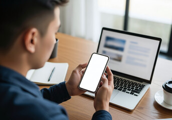 Man holding a smartphone in front of a laptop computer on a wooden desk, working remotely