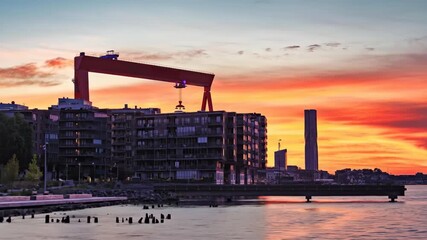 A waterfront cityscape illuminated by a vivid sunset with a massive crane rising over the harbor - Powered by Adobe