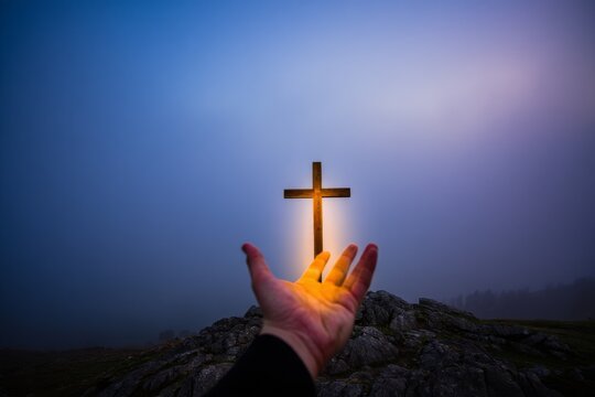 A hand holding up a glowing golden cross, radiating light against a dark, spiritual sky. Symbolic of faith, hope, divine light, and spirituality.
