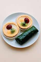 Delicipus fruit tarts and wrapped traditional snack on a white plate

A white plate holds two fruit tarts topped with fresh kiwi, grape, and peach pieces, alongside a traditional indonesia snack wrapp