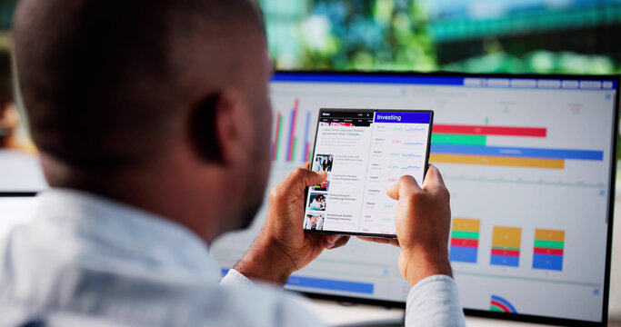 Young African American Man Trading Stocks On Foldable Phone Screen At Desk