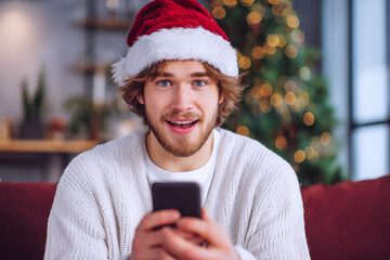 Close up portrait of joyful Caucasian handsome man in santa hat sitting in cozy room near xmas tree typing on smartphone with surprised face win christmas lottery. Happy New Year and Merry Christmas!
