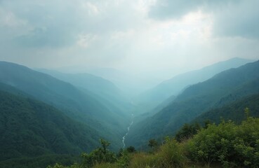Obraz premium Mountain valley with river flowing through green slopes under hazy sky. Layers of blue mountains recede into distance. Tall grass and foliage in foreground.