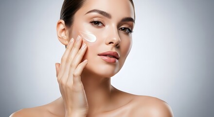 Beauty woman applying moisturizing face cream to cheek; natural makeup, healthy glowing skin, soft studio lighting, grey gradient background. clean skincare and cosmetics concept.