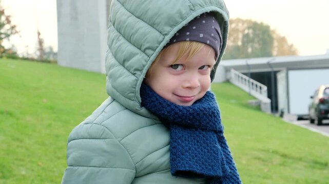 A four-year-old boy in a green jacket and blue scarf looks into the camera with a mischievous smile. Perfect 4K video for family, childhood, and lifestyle content.