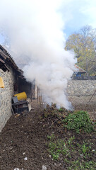 Fototapeta premium Smoke from burning leaves in the garden of a house in autumn. A bonfire burning between 2 stone houses. A bonfire smoking next to a stone fence.