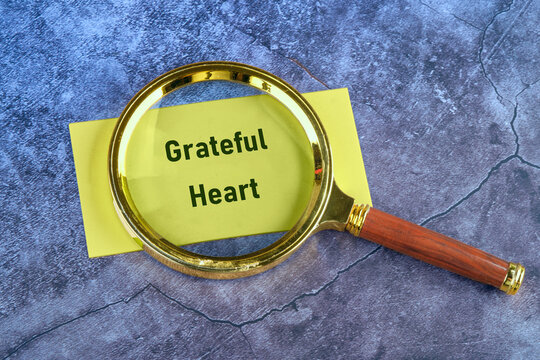 A magnifying glass highlights a yellow card displaying the words Grateful Heart, resting on a gray surface with cracks. It shows a reminder to be thankful
