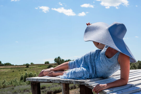 Young beautiful sexy woman in big hat lying on wooden bridge over lavada flowers field on sunny day against sky. Recreation. Vacation. Tourism. Enjoyment. Background for text.