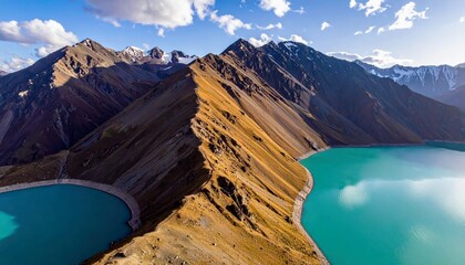A stunning aerial view of a vibrant turquoise lake curving around a sharp, golden mountain ridge, with snow-capped peaks in the distance under a clear blue sky.
