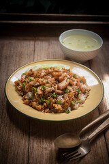 Delicious Thai-style fried chicken with rice, topped with crispy garlic and herbs, served with a bowl of clear soup on a wooden table. Authentic homemade Asian meal, beautifully presented for traditio