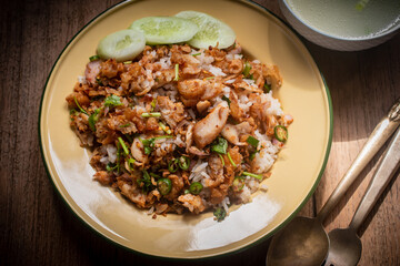 Delicious Thai-style fried chicken with rice, topped with crispy garlic and herbs, served with a bowl of clear soup on a wooden table. Authentic homemade Asian meal, beautifully presented for traditio