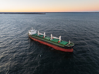 Naklejka premium High-angle aerial drone shot of a massive red bulk carrier cargo ship anchored in the tranquil Baltic Sea during golden hour.