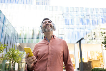 Happy mature man wearing headphones listening to music with phone app in the city.