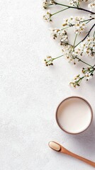 A close-up shot of delicate white blossoms and a bowl of creamy liquid with a wooden spoon on a textured white surface.
