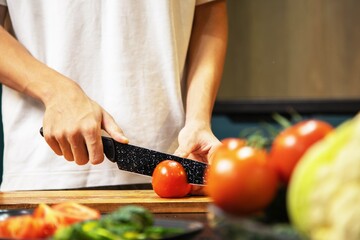 A person slicing a fresh tomato on a wooden cutting board while preparing a salad in the kitchen. Fresh vegetables and fruits lie nearby, creating a healthy cooking scene.