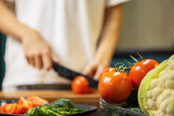 A person slicing a fresh tomato on a wooden cutting board while preparing a salad in the kitchen. Fresh vegetables and fruits lie nearby, creating a healthy cooking scene.